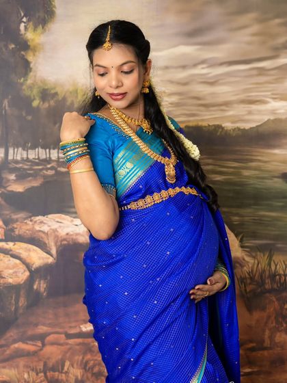 A close-up portrait celebrating motherhood with elegance. The focus is on the mom-to-be's serene expression, beautiful jewelry, and the rich blue of her traditional saree.