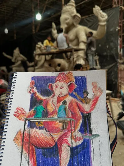 My coloured pencil sketch held up against the real-life scene at the Ganpati idol workshop. This shows how I translate the large-scale scene onto my sketchbook page.