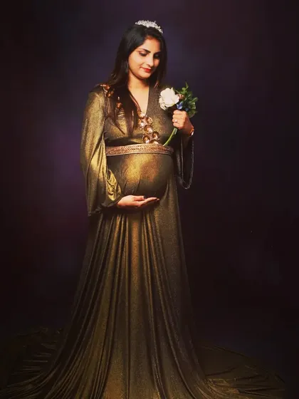 A classic studio maternity portrait. The mom-to-be is in a shimmering golden gown, holding a single white flower.
