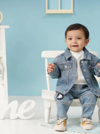 A stylish baby boy in a denim jacket poses for his first birthday portrait, sitting on a small white chair in a blue-themed studio set.