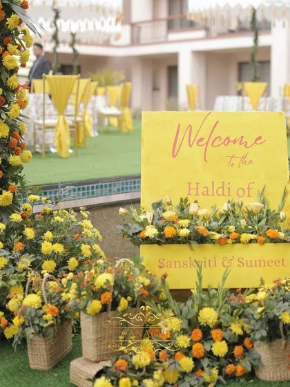 A bright and cheerful welcome setup for a Haldi, with a yellow sign nestled in a bed of marigolds and other yellow blooms in cane baskets.