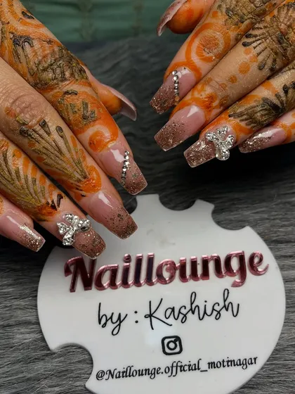 A close-up of a stunning bridal set against the client's mehendi. These long nails feature a glittery brown ombre effect, accented with sparkling 3D bow charms and rhinestones.