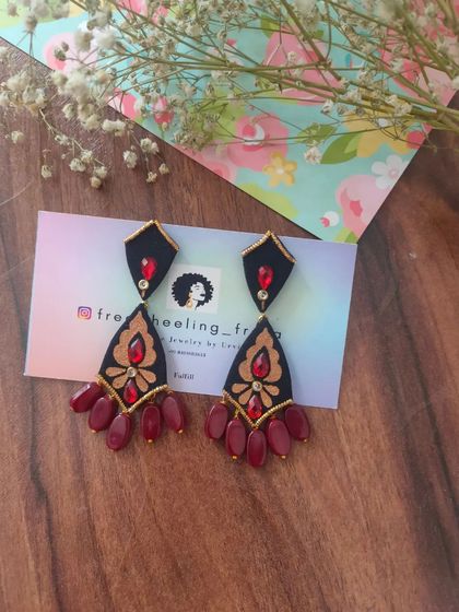 A pair of elegant, hand-painted chandelier earrings. The black fabric base is adorned with golden and red painted details and finished with maroon bead drops.