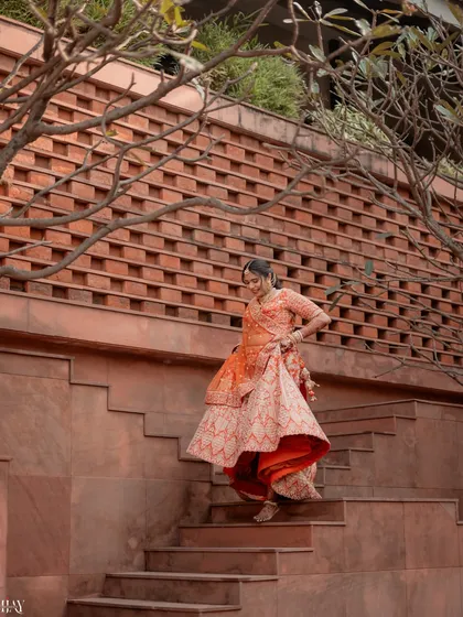 A candid shot of the bride descending the stairs, her orange lehenga in beautiful motion. This photo highlights the grace and elegance of the bridal outfit against the textured architectural backdrop.