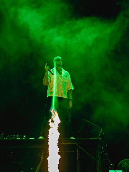 A column of fire shoots up from the stage as DJ Snake commands the crowd, surrounded by green smoke.