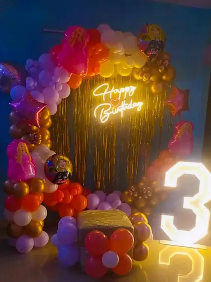 A Barbie-themed decoration for a 3rd birthday, using a colorful balloon arch, a gold fringe curtain, and a light-up number.