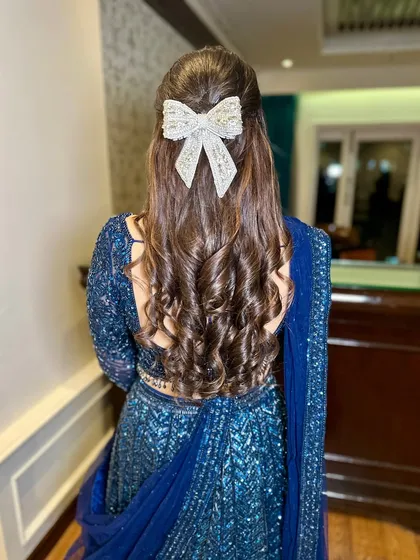 A modern hairstyle with soft curls and a chic bow, perfect for a Sangeet or reception look.