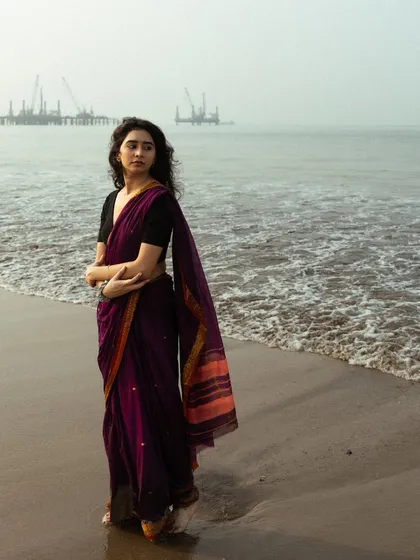 A full-length shot showing the model standing confidently on the shore, with the industrial pier adding a gritty contrast to the elegant saree.