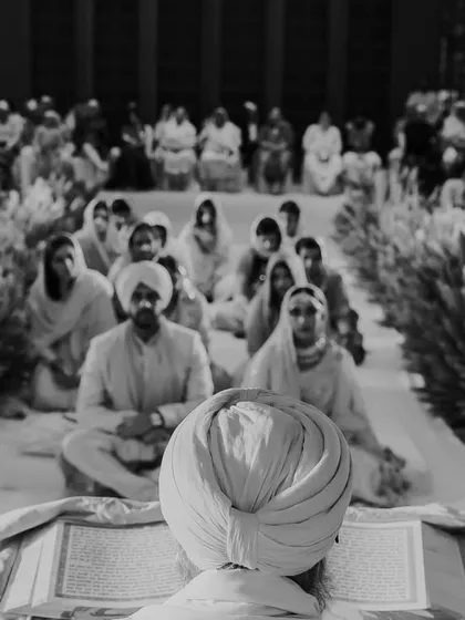 A timeless black and white photograph of the Anand Karaj. This view from behind the Granthi focuses on the couple and their families, seated in prayer, capturing the reverence and solemnity of the sacred union.
