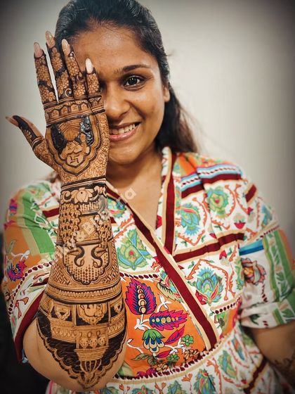 A happy bride showing off her full arm mehndi. The design is a beautiful blend of traditional figures, including a Ganesh portrait, and personalized initials.