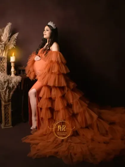 A classic and elegant solo portrait. The mother-to-be, wearing a crown and a tiered orange gown, poses gracefully in a candlelit, rustic studio setting.