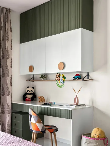 The study area in this kids' room is designed to be both fun and functional. It features two-tone cabinets with olive green fluted panels and white doors, a geometric print chair, and plenty of space for books and toys.