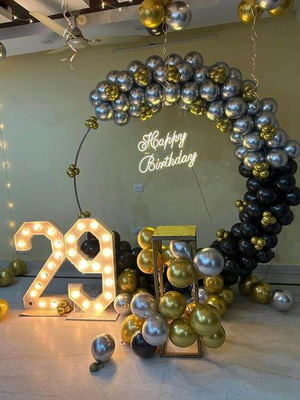 A modern and stylish 29th birthday setup. The decor features a circular balloon frame in black, silver, and gold, a 'Happy Birthday' neon sign, and large marquee numbers.
