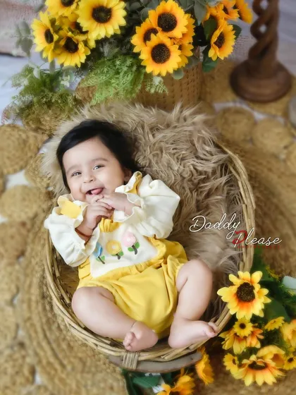 A joyful baby girl with a big smile, sitting in a rustic basket surrounded by bright sunflowers. The natural textures and cheerful flowers create a warm and happy portrait.