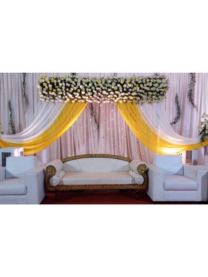 A bright and cheerful stage design ideal for a Haldi or Sangeet ceremony. The combination of yellow and white drapes, a dense floral header, and modern white sofas creates a fresh and inviting look.