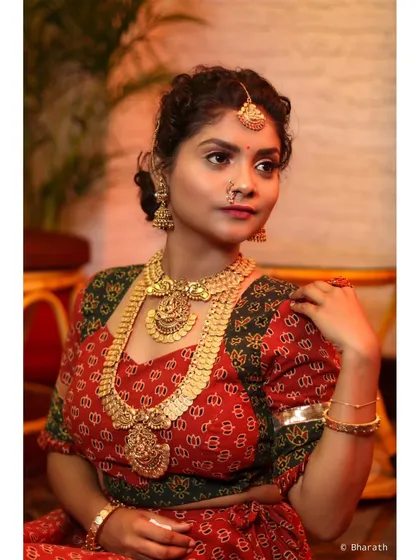 A three-quarter view of a model in a traditional red and green outfit. The pose showcases the full set of temple jewelry against the patterned fabric.