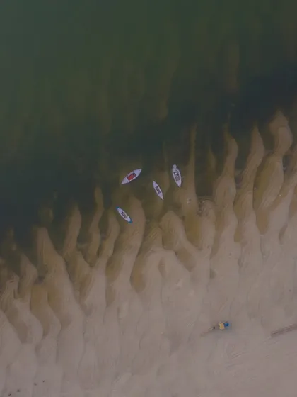 A top-down view of kayaks resting on the sandy banks of the Ganga in Varanasi. The patterns in the sand and the clear water create a beautiful, minimalist composition.