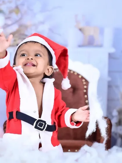 A baby dressed as Santa Claus, waving happily amidst a snowy setup.