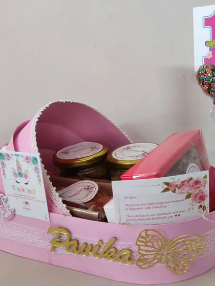 A single personalized cradle hamper for 'Pavika's' first birthday. It includes jars of treats, a chocolate lollipop, and custom unicorn-themed cards, all nestled in the beautiful pink cradle.