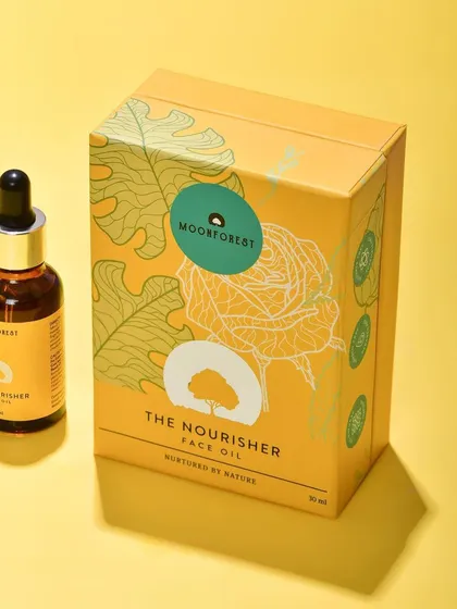 A bright and cheerful shot of Moon Forest's "The Nourisher" face oil next to its box against a yellow background. This is a great e-commerce shot that shows both the product and its packaging.