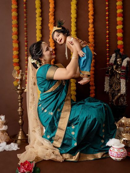 A mother in a beautiful teal saree lifts her laughing baby, who is dressed as Krishna, creating a vibrant and happy portrait.