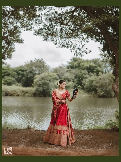 A beautiful, full-length portrait of the bride by the water. The natural landscape provides a serene and timeless backdrop for her traditional red and gold lehenga.