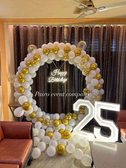 A clean and classic 25th birthday or anniversary setup in a living room, with a white and gold balloon ring, "Happy Birthday" neon sign, and "25" marquee numbers.