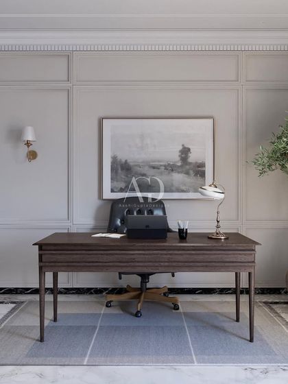 Soft minimalism in a home office. The design uses a simple, clean-lined desk and a modern leather chair, set against a backdrop of classic wall paneling. An olive tree adds a touch of nature and softness to the formal space.