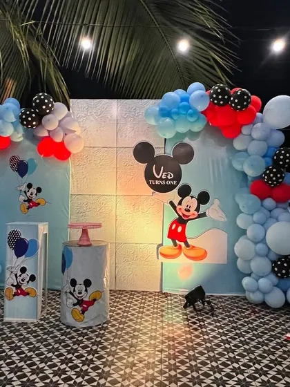 A classic Mickey Mouse themed first birthday party set up on a terrace. The decor includes character cutouts, themed pedestals, and a balloon arch in Mickey's signature red, blue, and black colors.