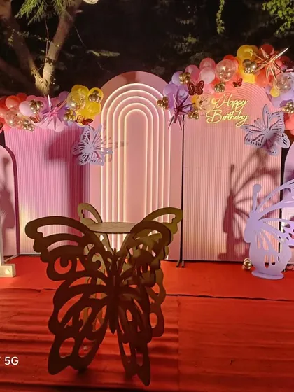 This is a beautiful butterfly-themed birthday setup for an outdoor evening party. It features pink arched backdrops, a custom butterfly-shaped table, and a colorful balloon garland.