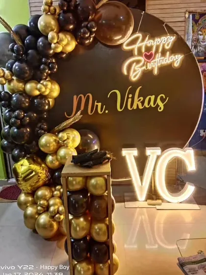 A sophisticated birthday setup for Mr. Vikas. The black circular backdrop is accented with a black and gold balloon arch and his initials in marquee letters.