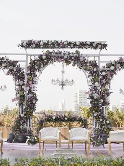 A beautiful daytime view of Reeya and Parth's rooftop wedding mandap. The structure of white arches is adorned with lush lavender and pink florals, creating a romantic garden feel against the city skyline.
