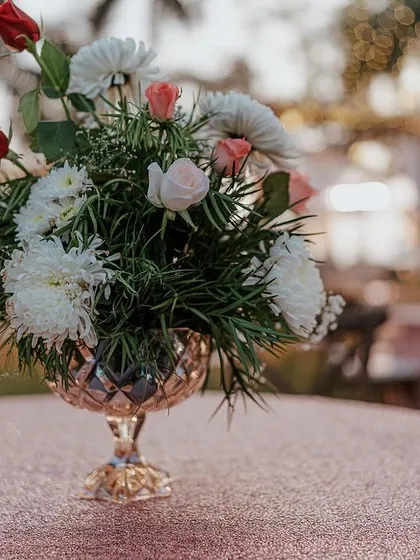 A close-up of a table centerpiece at an outdoor reception. The simple arrangement of roses and chrysanthemums in a crystal vase adds a touch of elegance to the decor.