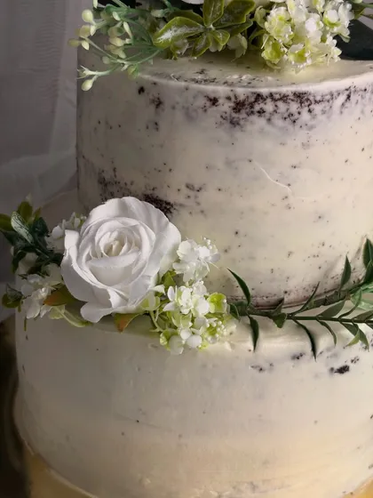 A detailed close-up of my rustic semi-naked cake. You can see the texture of the light buttercream coating over the dark cake, complemented by the delicate arrangement of food-safe white flowers and greenery.