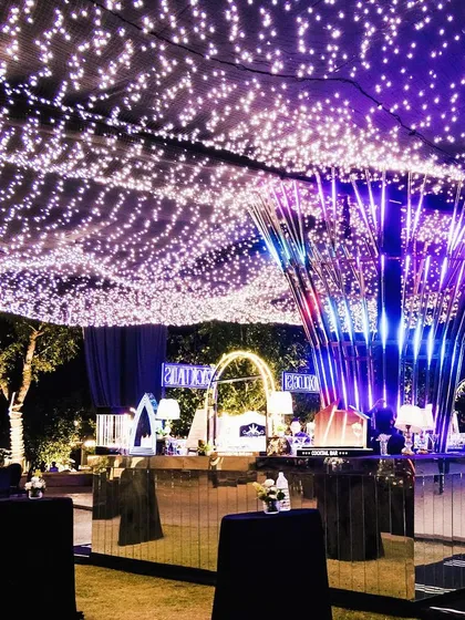 A glamorous outdoor Sangeet setup featuring a ceiling of fairy lights, a modern light sculpture, and mirrored bar tables, creating a vibrant and sophisticated party atmosphere.