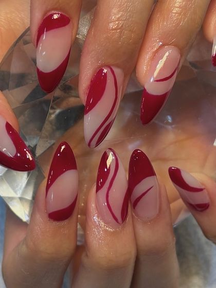Sassy and sweet cherry red nails. This abstract design features flowing red swirls on a sheer base, creating a dynamic and eye-catching look on almond-shaped extensions.