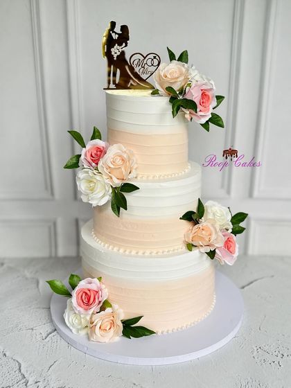 A classic three-tier wedding cake perfect for a grand celebration. The design features a soft peach ombre effect, adorned with bouquets of fresh roses and a romantic silhouette topper of the bride and groom.