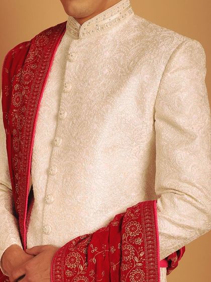 A close-up of the fine, self-patterned fabric of an ivory sherwani, paired with a richly embroidered red velvet stole. This highlights the luxurious textures we use in our creations.