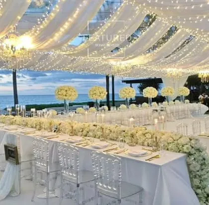 This all-white reception setup by the sea is the essence of elegance. I used white floral runners, transparent ghost chairs, and a ceiling of draped fabric with fairy lights to create a breathtaking, monochromatic look.