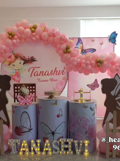 A sweet butterfly and fairy theme for a first birthday, using a circular backdrop and fairy silhouette cutouts. The decor includes butterfly-printed plinths and a pink balloon arch with gold accents.