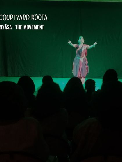 A solo Bharatanatyam performance of 'Vinyāsa - The Movement'. The dancer is captured mid-pose, with dramatic green lighting creating a powerful silhouette against the audience.