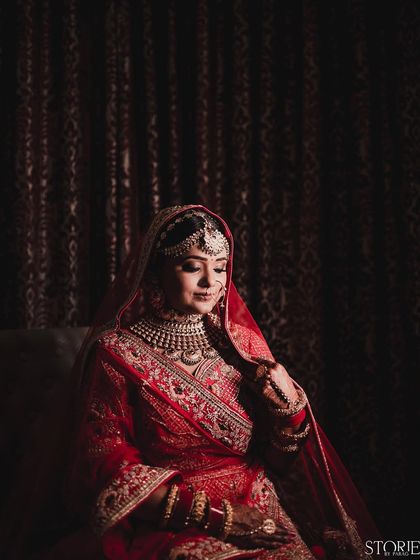 A classic, moody bridal portrait. The bride, dressed in a rich red lehenga, is seated in soft, dramatic light that highlights the intricate details of her attire and her serene expression.