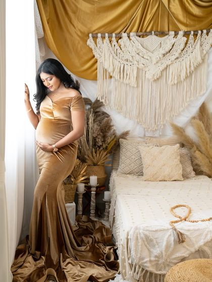 A solo portrait of an expecting mother in a golden velvet gown. She stands by a window in a boho-chic room, looking down at her bump with a gentle expression.