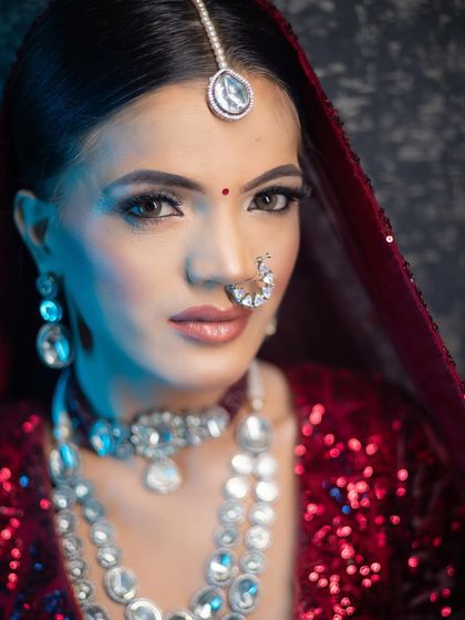 A striking close-up of a bridal look under dramatic blue lighting. This highlights the sharp, defined eye makeup, the contour, and the beautiful diamond jewelry against the red lehenga.