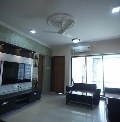 This living room features a TV unit with a green textured accent wall and an attached white marble pooja mandir. The layout is simple and functional, with a leatherette sofa set.