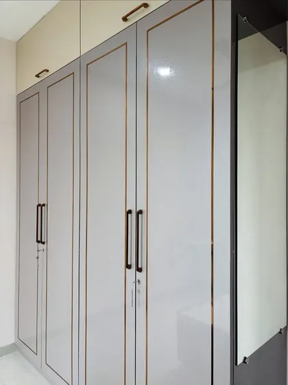 A close-up of a four-door hinged wardrobe with a light grey glossy finish and elegant gold trim. This design includes an attached dressing unit with a mirror, combining storage and functionality.