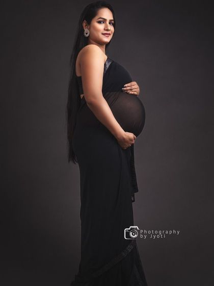 A classic and elegant maternity portrait in a sheer black saree against a dark grey background, highlighting the baby bump silhouette.