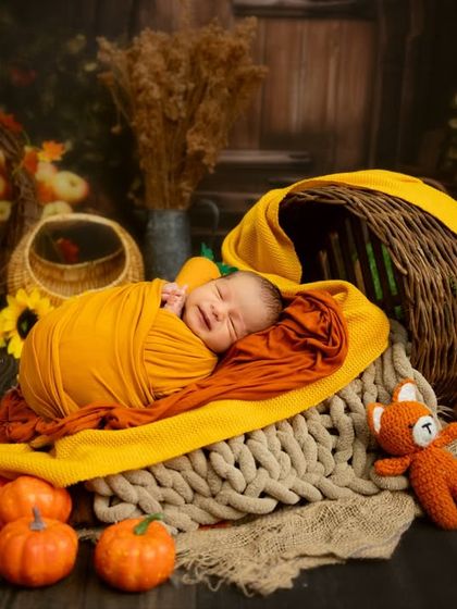 An autumn-themed newborn shoot full of warmth. The baby is swaddled in orange and yellow wraps, sleeping in a basket surrounded by miniature pumpkins and fall leaves.