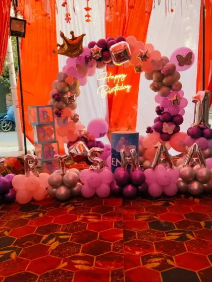 A grand first birthday setup for "Trishaan" in an outdoor tent setting. This decoration includes a circular balloon arch, the baby's name in foil letters, and a "Happy Birthday" neon sign.