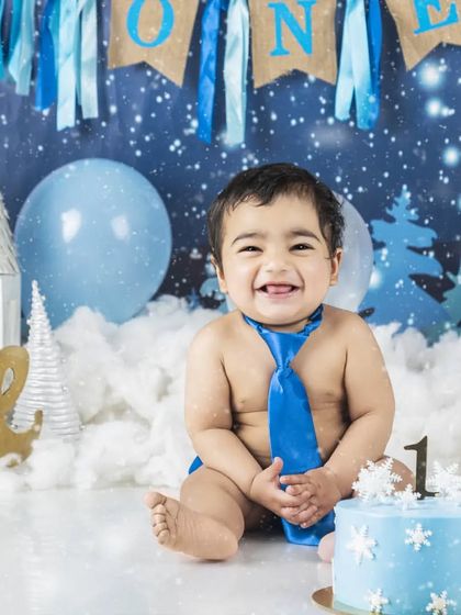 A big, happy smile from the birthday boy at his winter-themed cake smash. The blue tie and snowflake cake create a cool and fun first birthday photo.